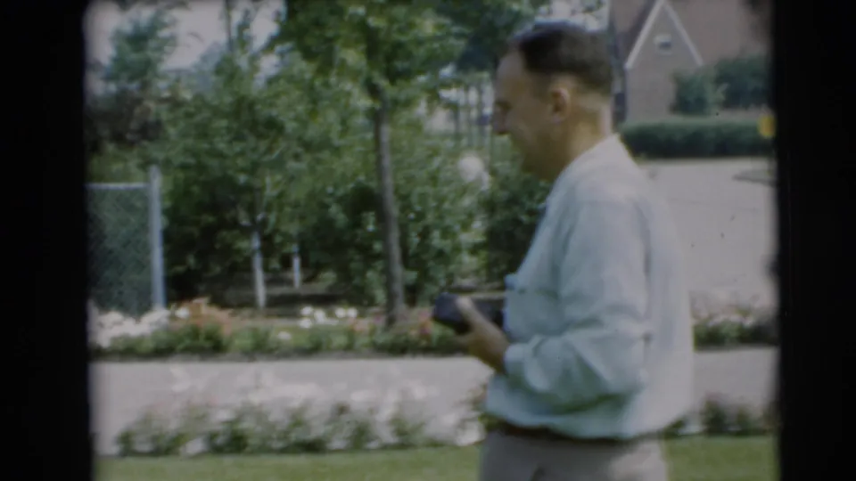A man in a light shirt walks through the 1954 Pennington yard scene holding a small dark object, with flowers and houses behind him.