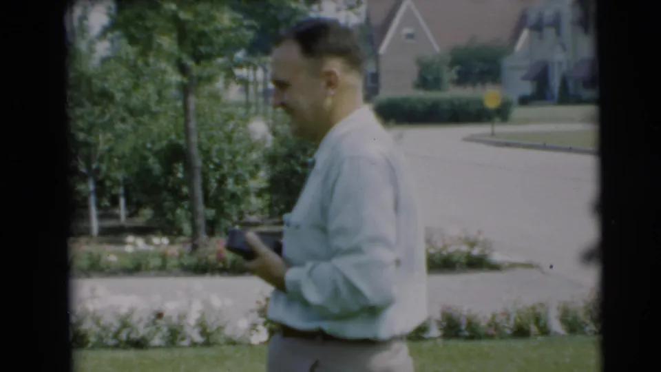 The man appears in profile in the Pennington yard, holding a small object with a flower bed, fence, street, and houses in the background.