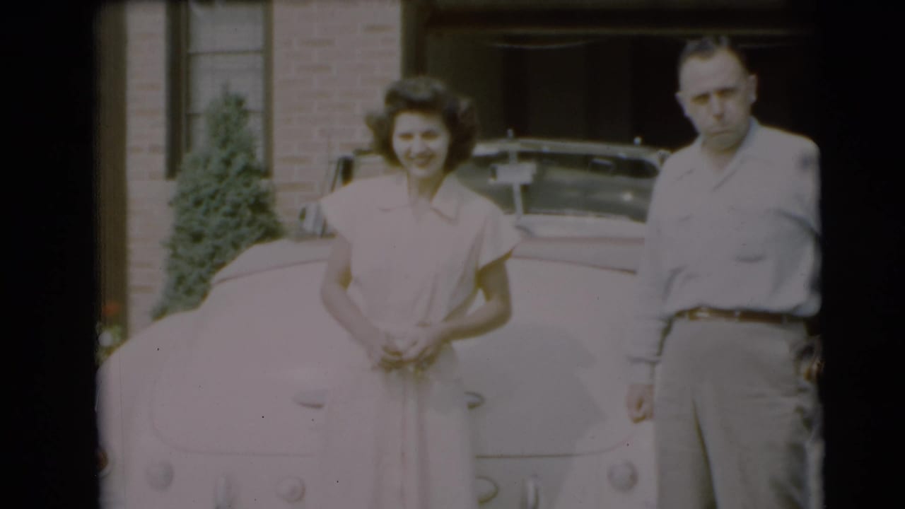 In Pennington, New Jersey, 1954, a young woman and man stand near a pale vintage car in front of a brick residence.