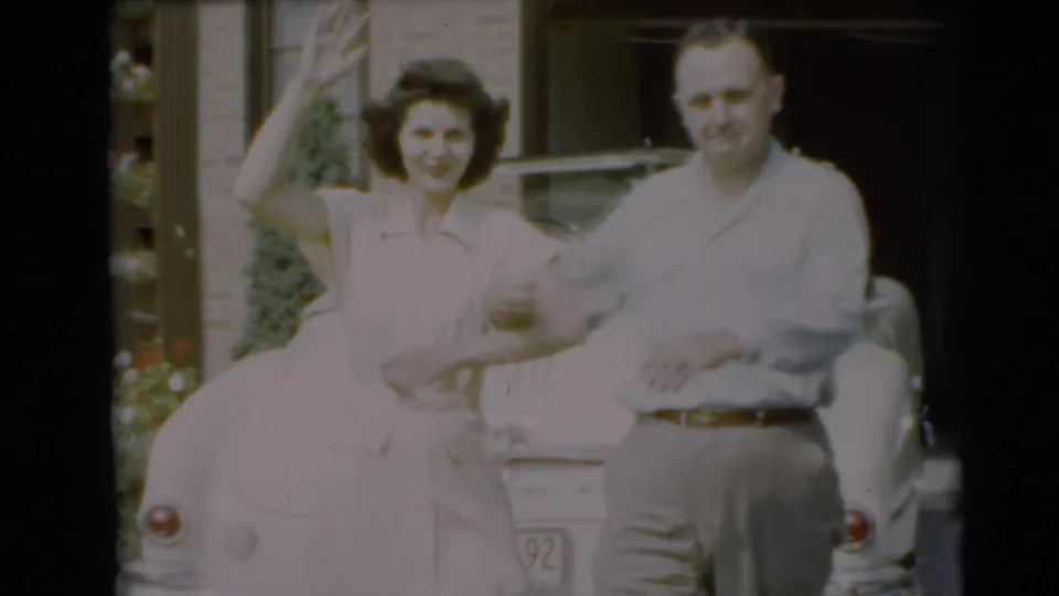 The woman waves toward the camera while standing with a man beside a car outside a Pennington brick home in 1954.