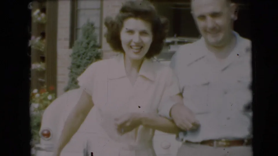 A smiling woman and man appear closer to the camera, with the vintage car and brick house still visible behind them.