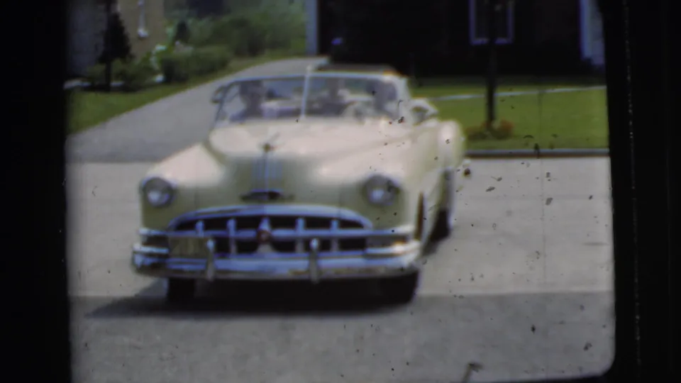The yellow convertible remains near the driveway entrance, with chrome trim, lawn, and a house exterior visible in soft 8mm focus.
