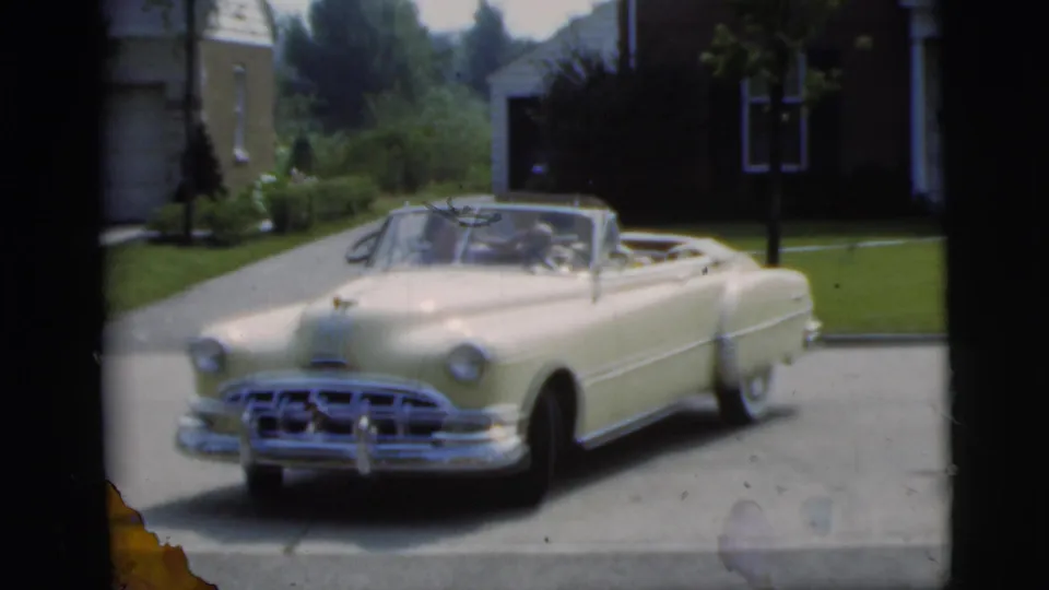 The yellow convertible is angled in the driveway in Pennington, New Jersey, with two occupants visible and a house exterior and lawn behind it.