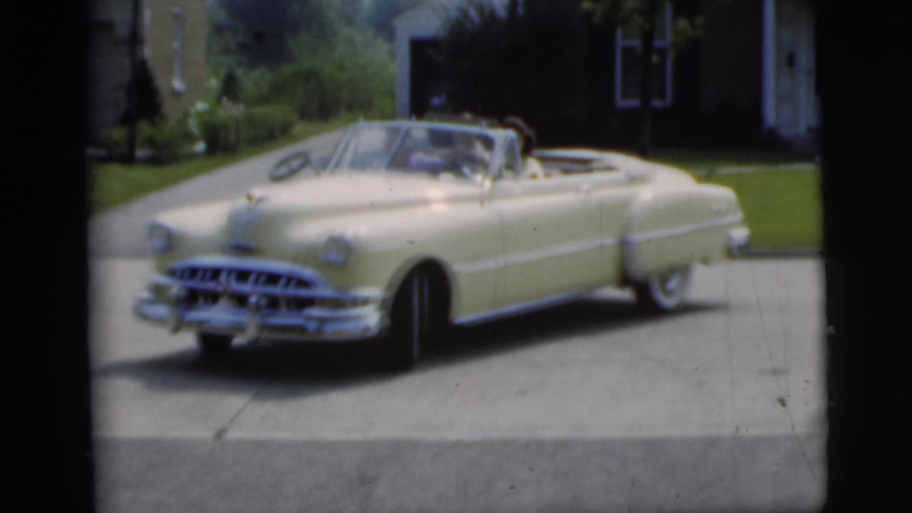 Cream 1950s convertible angled in a driveway near a brick house and lawn.