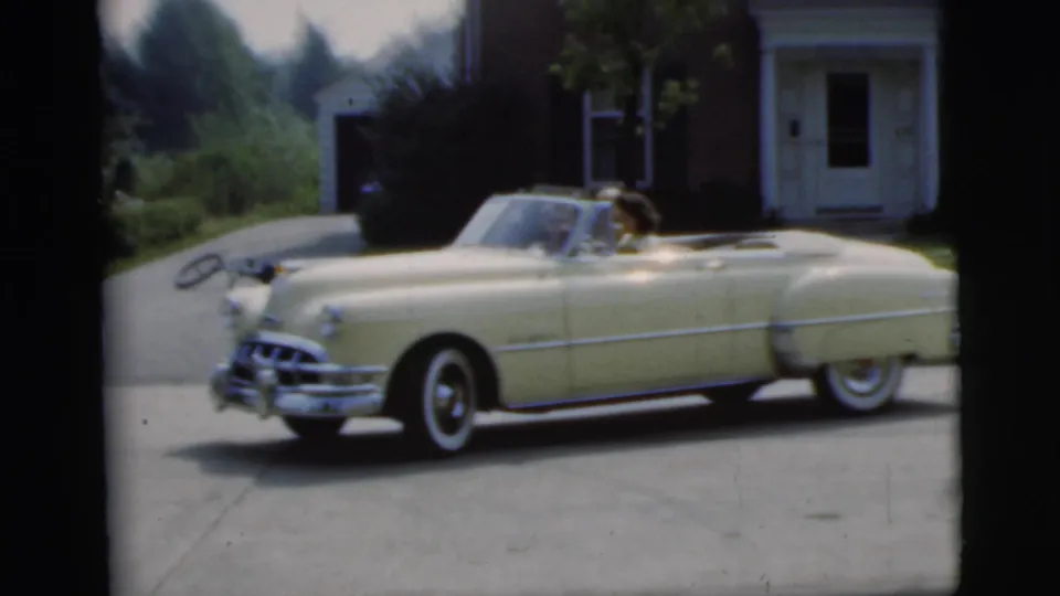 Side view of the cream convertible backing along a residential driveway in 1955 Pennington, with a passenger visible.