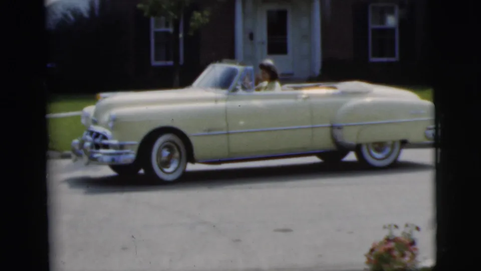 Cream 1950s convertible in side profile with a passenger visible, filmed outside a Pennington residential house in 1955.