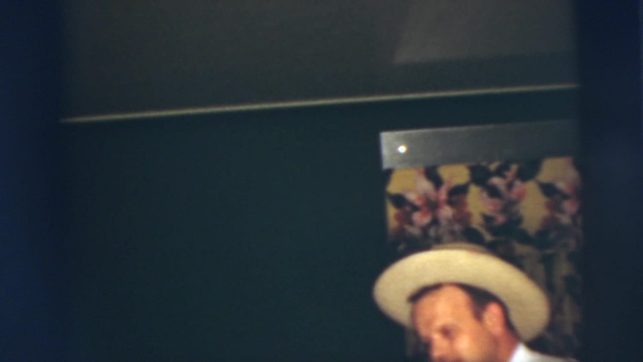In a 1955 Pennington, New Jersey home-movie frame, a young man in a light-colored hat stands partly cropped near floral wallpaper.