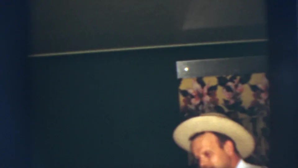 In a 1955 Pennington, New Jersey home-movie frame, a young man in a light-colored hat stands partly cropped near floral wallpaper.