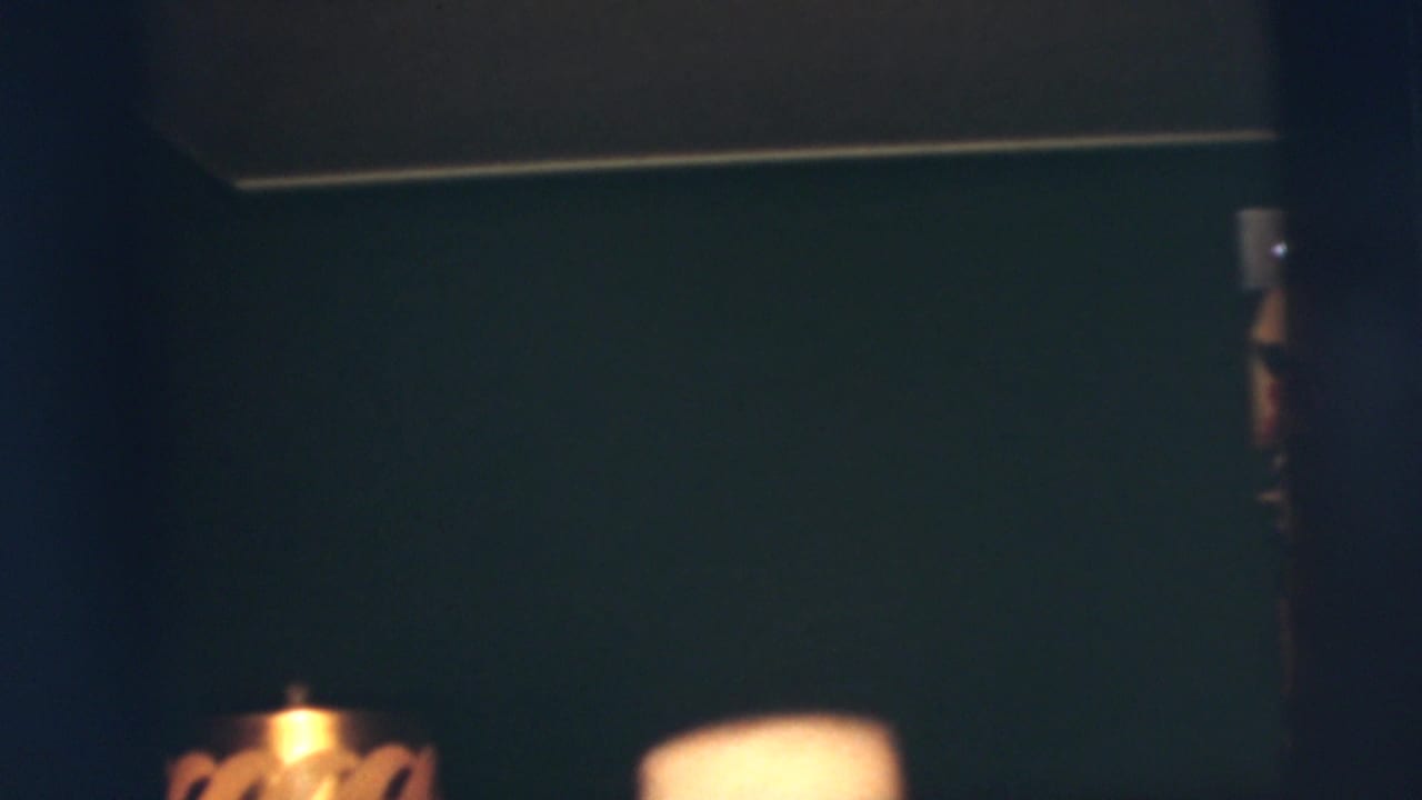 Low-light 1955 home-movie frame showing a dark domestic wall, lamp shade edge and partial adult face at the right.