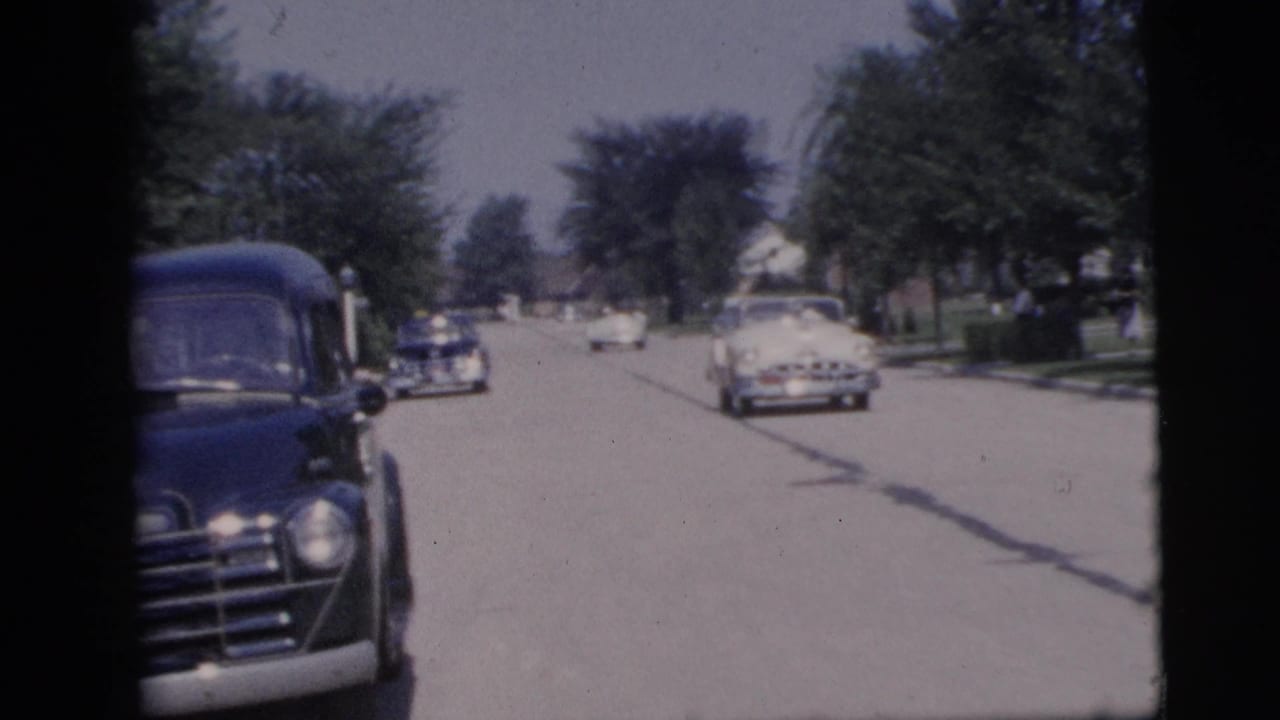 1955 Pennington, New Jersey home-movie frame showing a dark sedan near the camera and cars moving along a sunny residential street.