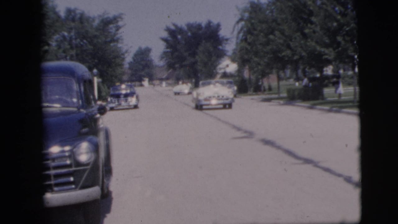 A dark mid-century car passes close to the lens on a sunny Pennington street as other vehicles continue along the road.