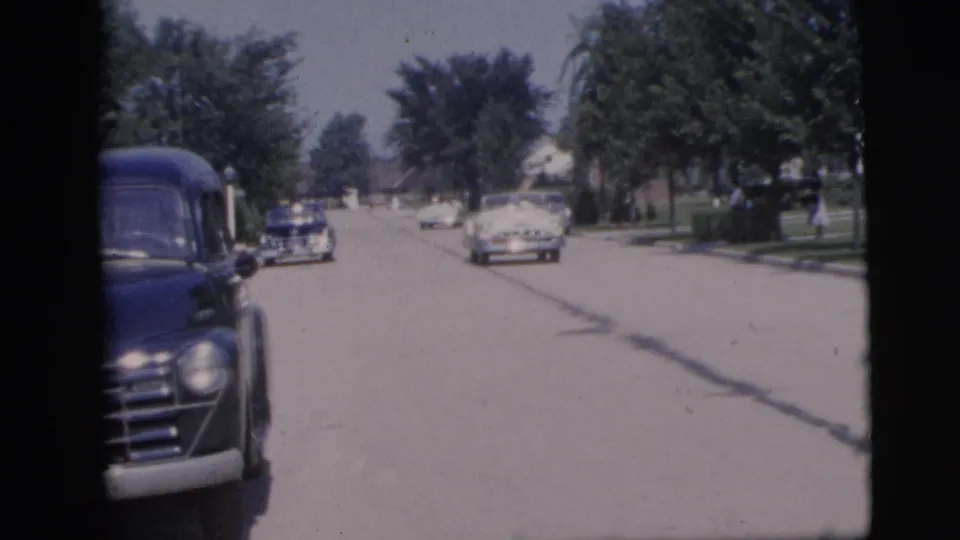 A dark mid-century car passes close to the lens on a sunny Pennington street as other vehicles continue along the road.