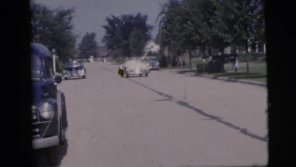 Mid-1950s Pennington street view with a dark car at the edge of frame and several cars moving through a leafy neighborhood.