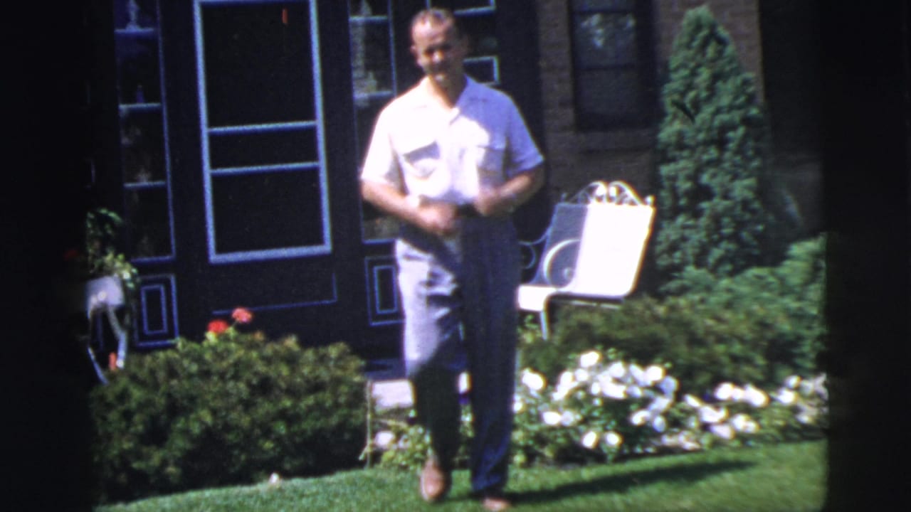 Adult man in a light shirt and trousers stands in a Pennington, New Jersey garden in 1955, with flowers, shrubs, and a white chair behind him.
