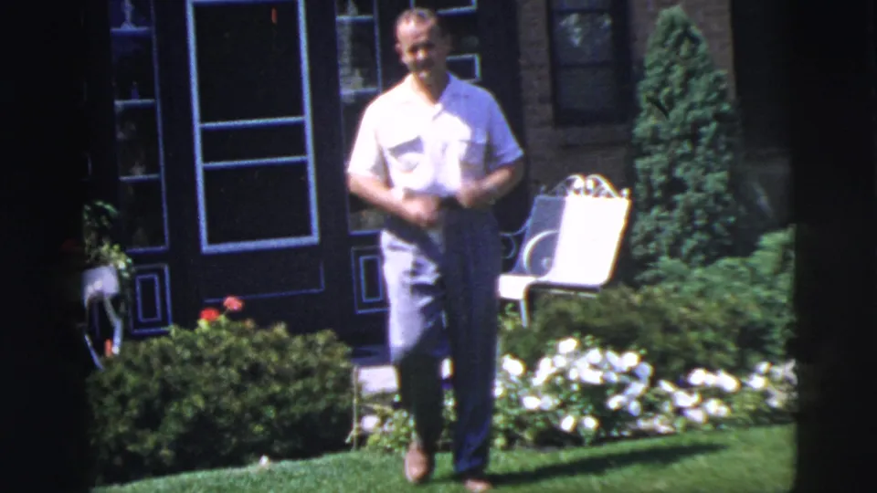 Adult man in a light shirt and trousers stands in a Pennington, New Jersey garden in 1955, with flowers, shrubs, and a white chair behind him.