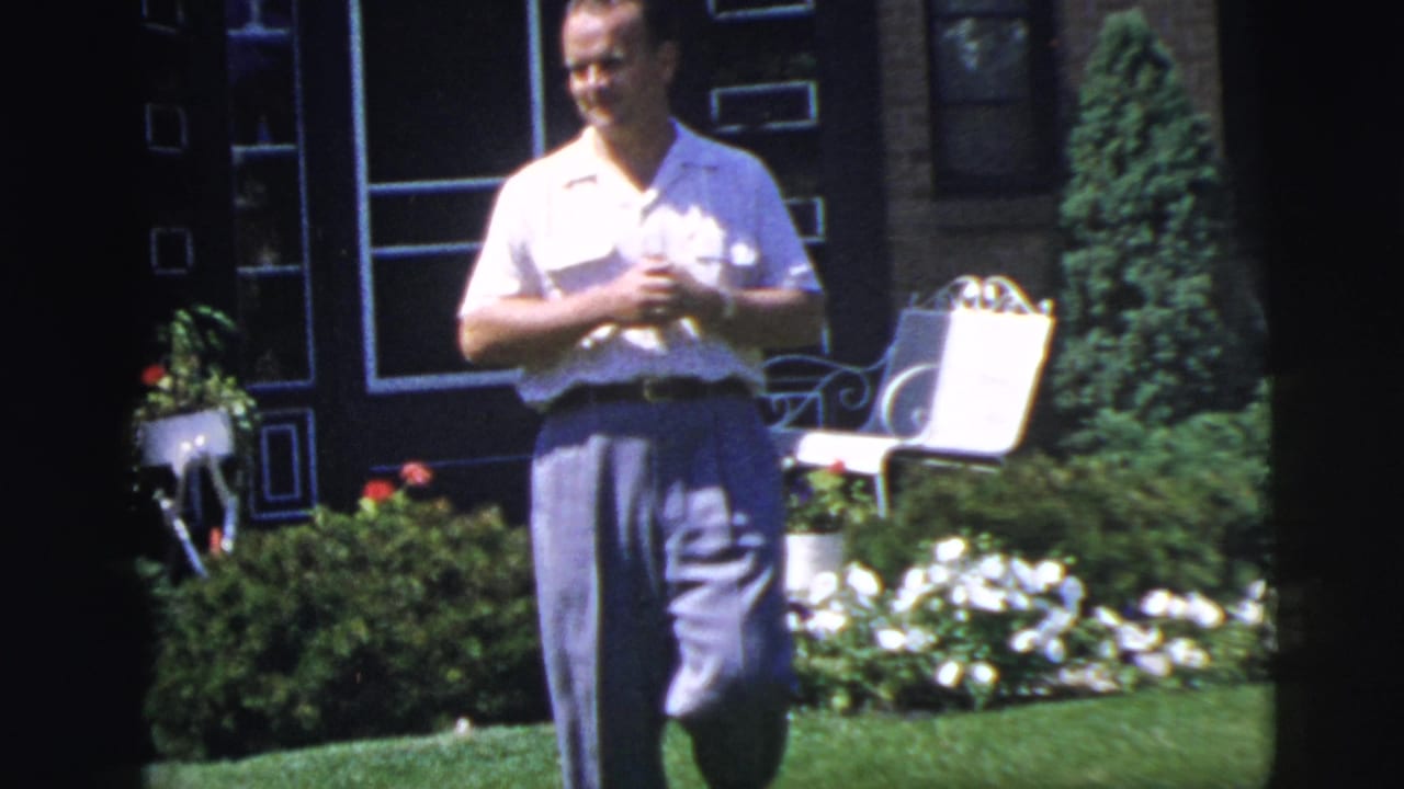 Man walking toward the camera across a lawn with garden flowers behind him.