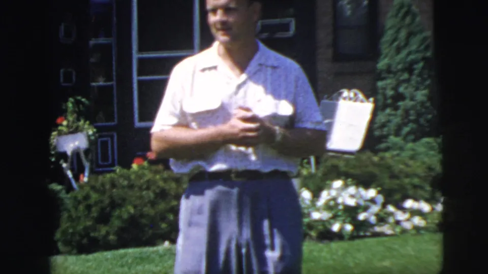 Closer 8mm frame of the man in a light shirt and blue-gray trousers holding a small object, with flowers and a white garden chair behind him.