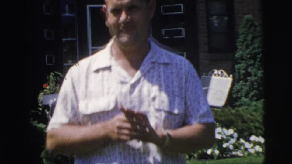 The man fills much of the frame in the Pennington garden, smiling toward the camera with the house entrance, flowers, and garden seating in the background.