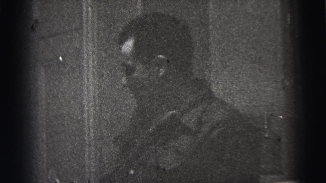 Grainy 1956 New York City home-movie frame of a man in profile, partly hidden by darkness near a wall or doorway.