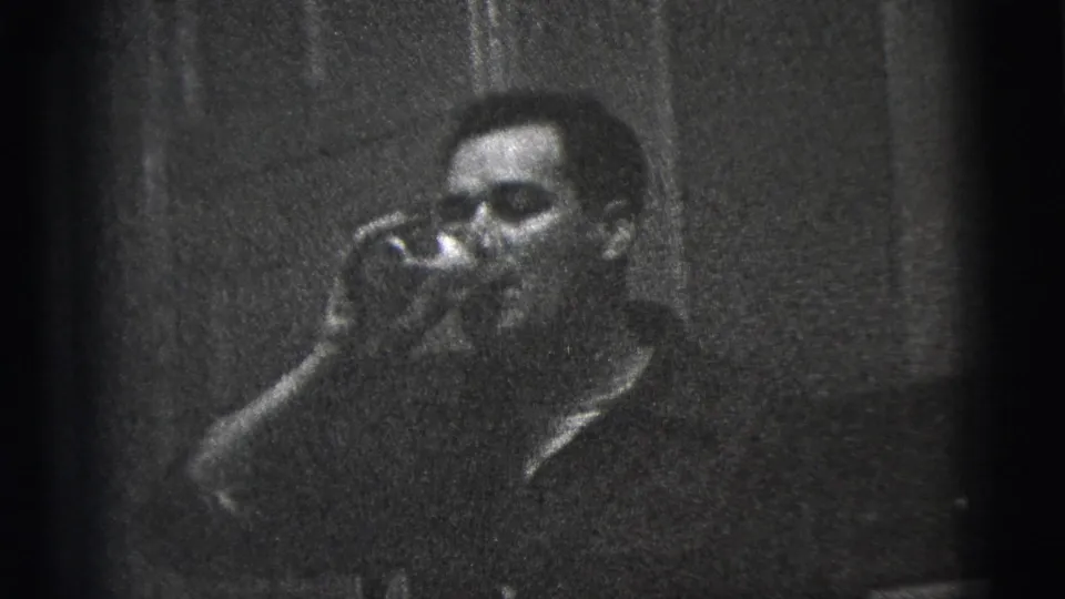 In 1956 New York City home-movie footage, a man in a dark collared shirt raises a glass to drink inside a dim private kitchen or interior.