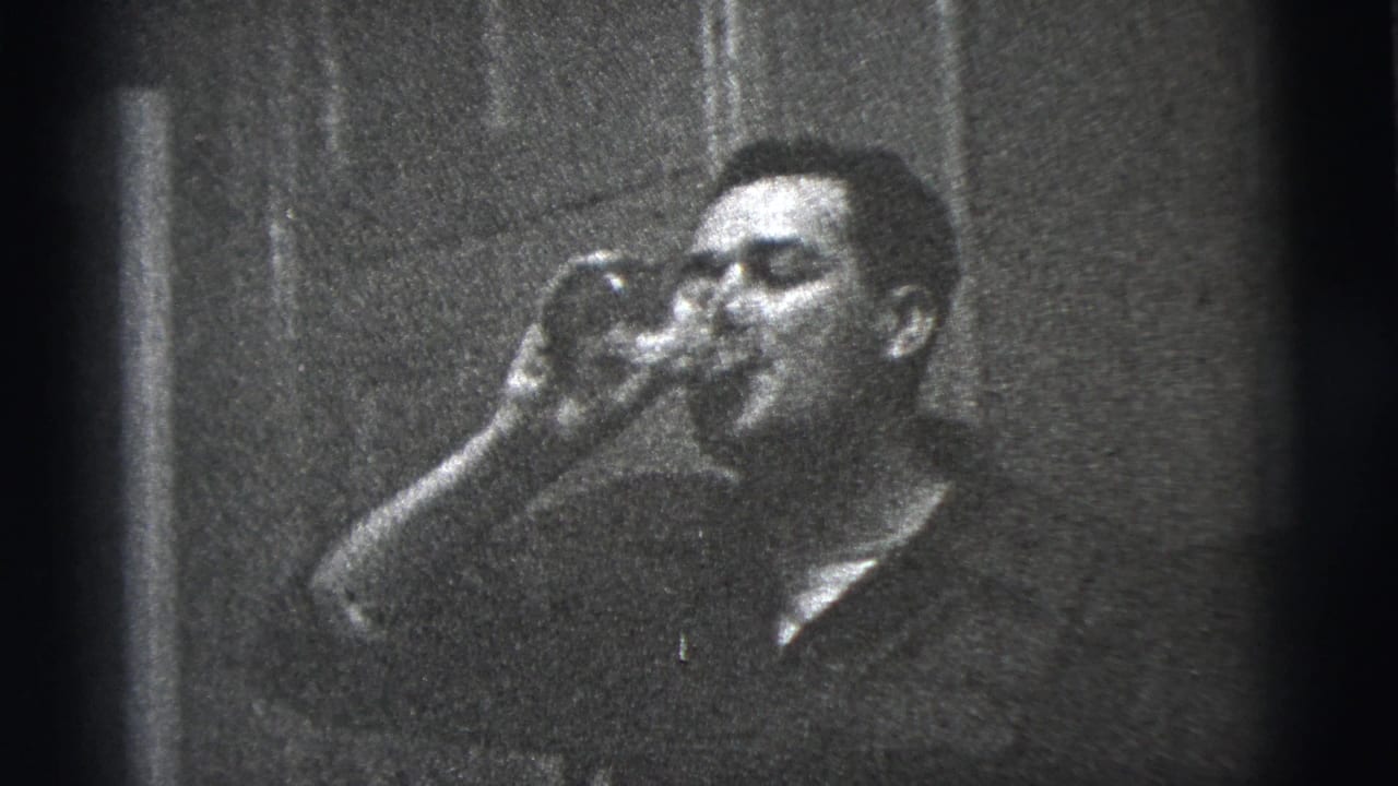 The man continues drinking from a glass in a grainy 1956 New York City interior, with a plain wall or door surface behind him.