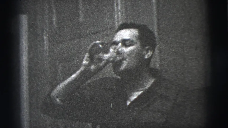 The man continues drinking from a glass in a grainy 1956 New York City interior, with a plain wall or door surface behind him.