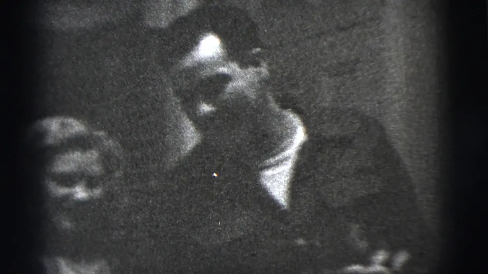 A smiling woman appears beside the man in the grainy black-and-white 1956 New York City home-movie interior.