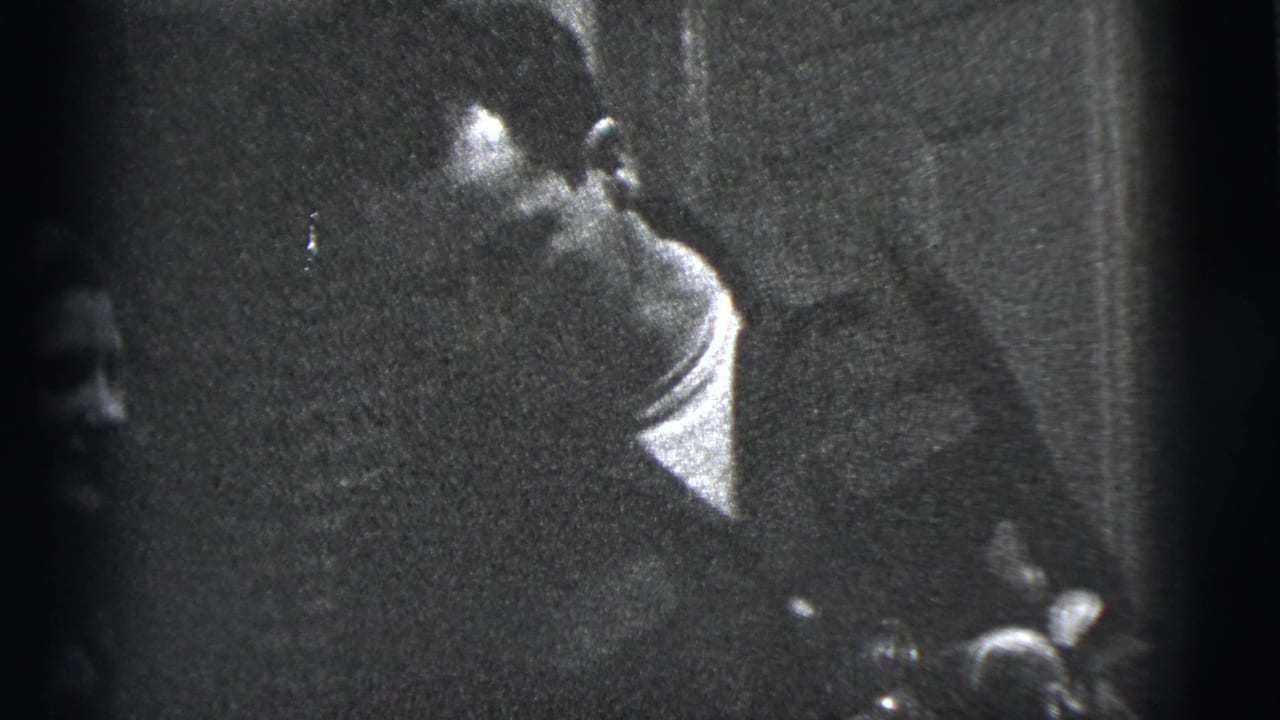 In a 1956 New York City home-movie frame, a seated man turns in a dim room while a woman is partly visible nearby.