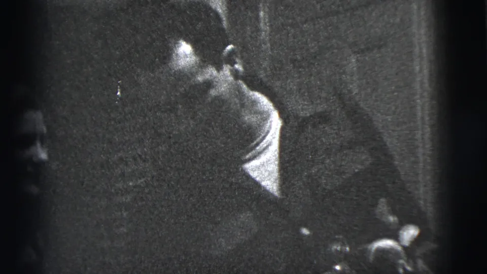 In a 1956 New York City home-movie frame, a seated man turns in a dim room while a woman is partly visible nearby.