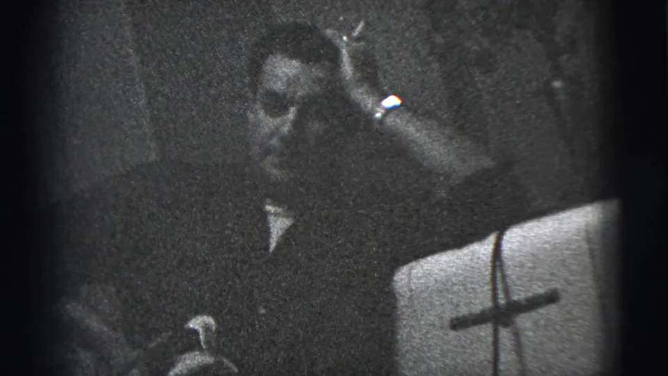 A seated man in a dark shirt raises a cigarette near his face in a dim 1956 New York City interior.