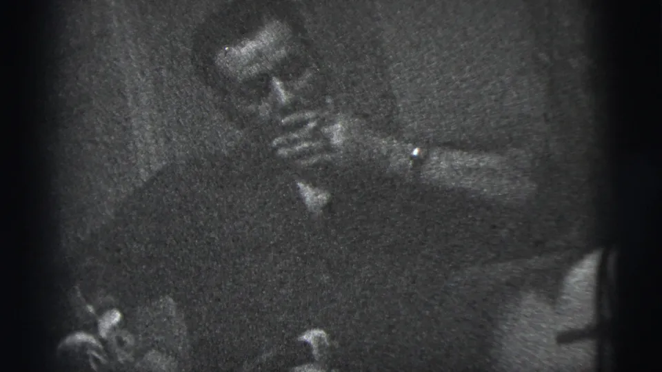 The man continues smoking while seated in a dark private interior, captured in grainy 1956 New York City 8mm footage.