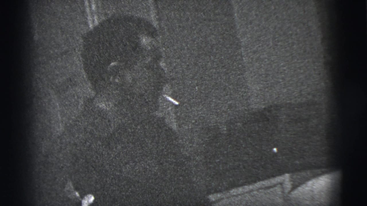 Seated man in a dark suit holding a cigarette in a dim interior.