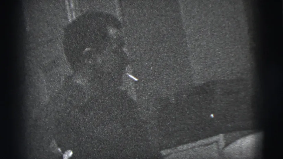 A man in a dark suit appears seated indoors in 1956 New York City home-movie footage, holding or smoking a cigarette.