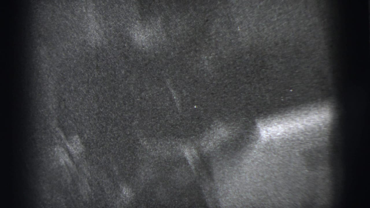 A dim 1950s home-movie frame shows the seated man partly obscured by shadow and close handheld framing indoors.