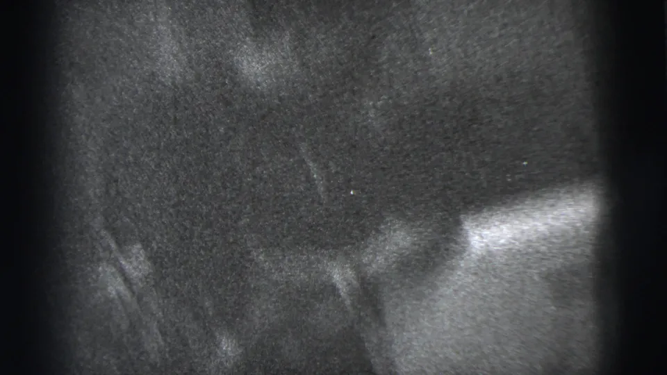 A dim 1950s home-movie frame shows the seated man partly obscured by shadow and close handheld framing indoors.