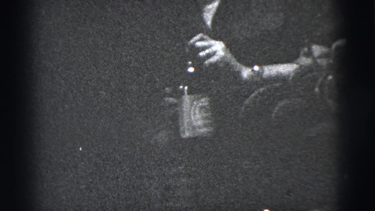 Grainy close view of a hand holding a bottle and cigarette near a dark sleeve.