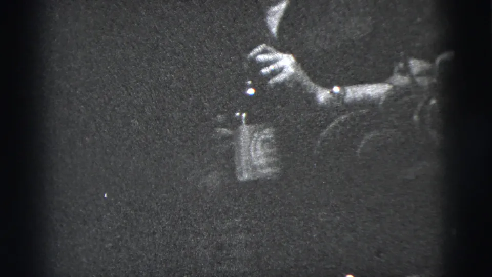 1956 New York City home-movie frame showing a hand with a cigarette and bottle, with a wristwatch and dark clothing visible in low light.