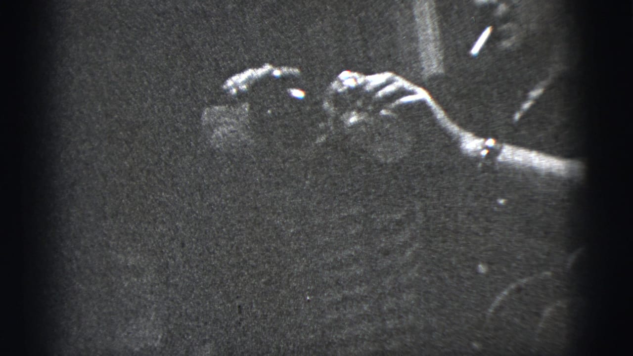 Black-and-white 1956 New York City home-movie frame showing hands with a wristwatch handling a drink in a dark lounge-like interior.