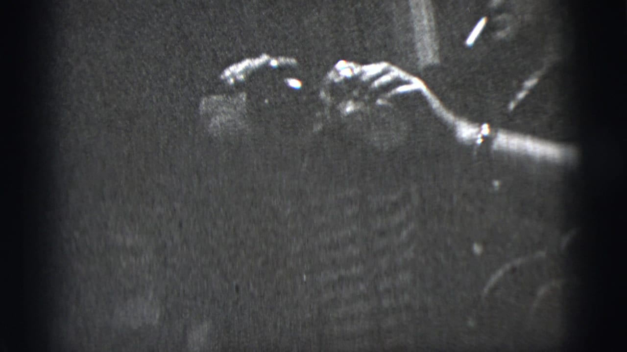 Black-and-white frame from the supplied 1956 New York City clip with hands, a wristwatch, and a partially visible person in low light.