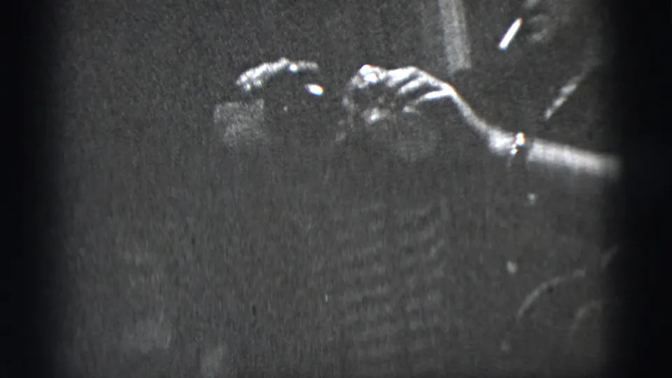Black-and-white frame from the supplied 1956 New York City clip with hands, a wristwatch, and a partially visible person in low light.