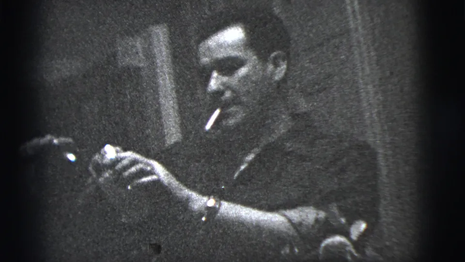 Black-and-white 1956 New York City home-movie frame showing a man with a cigarette holding a drink in a lounge-like interior.