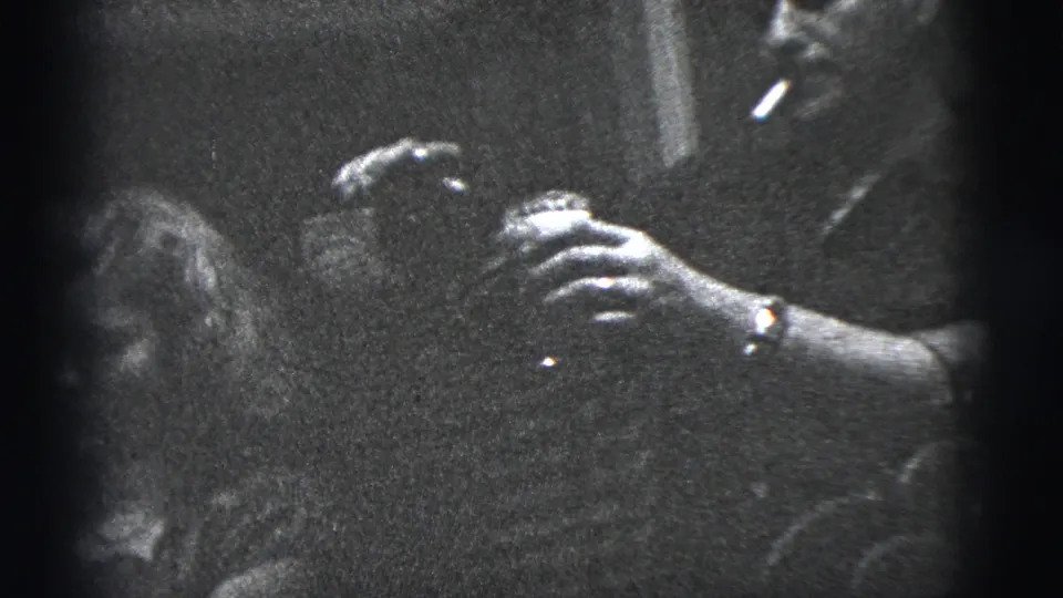Black-and-white frame from a 1956 New York City home movie showing hands with a drink and a nearby seated person in a dark lounge interior.