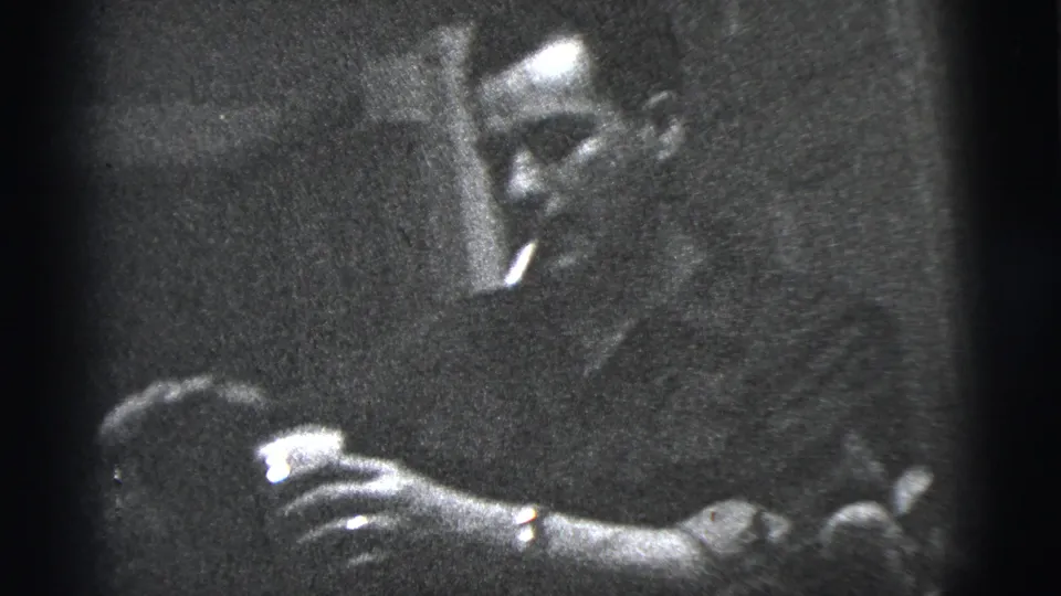 In 1956 New York City home-movie footage, a man and woman stand close together while hands reach toward a small bag or object.