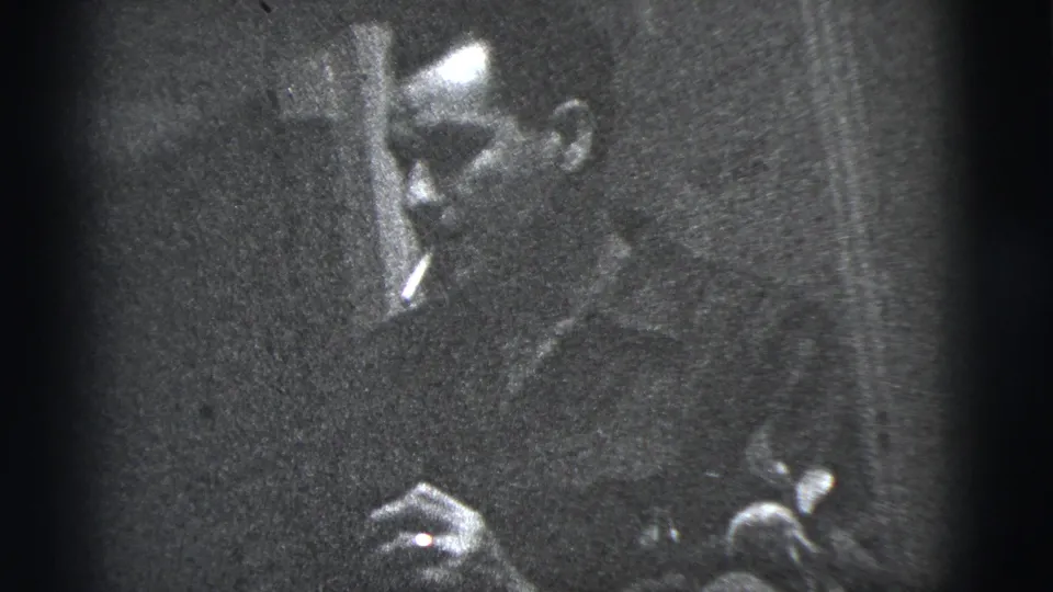 A man appears in close-up in 1956 New York City home-movie footage, with a hand and small object visible lower in the frame.