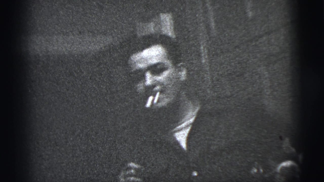 1956 New York City home-movie frame of an adult man in a dark jacket holding a cigarette while facing the camera.