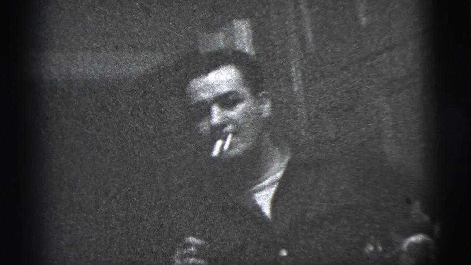 1956 New York City home-movie frame of an adult man in a dark jacket holding a cigarette while facing the camera.