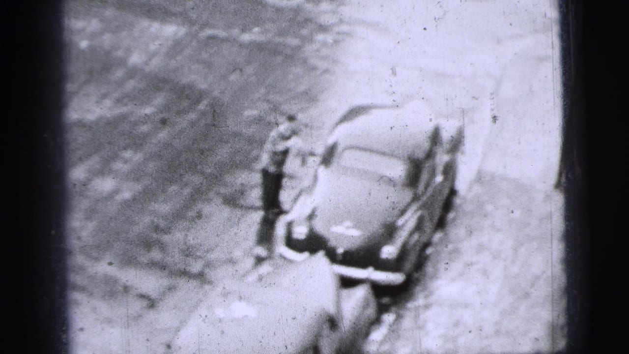 High-angle 1956 New York City 8mm frame showing a man standing beside a parked mid-century car at the curb.