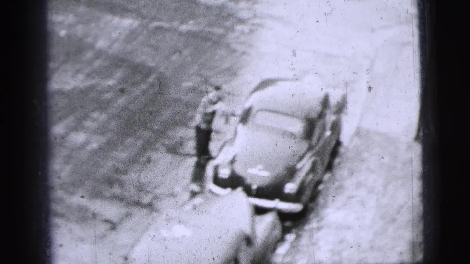 High-angle 1956 New York City 8mm frame showing a man standing beside a parked mid-century car at the curb.