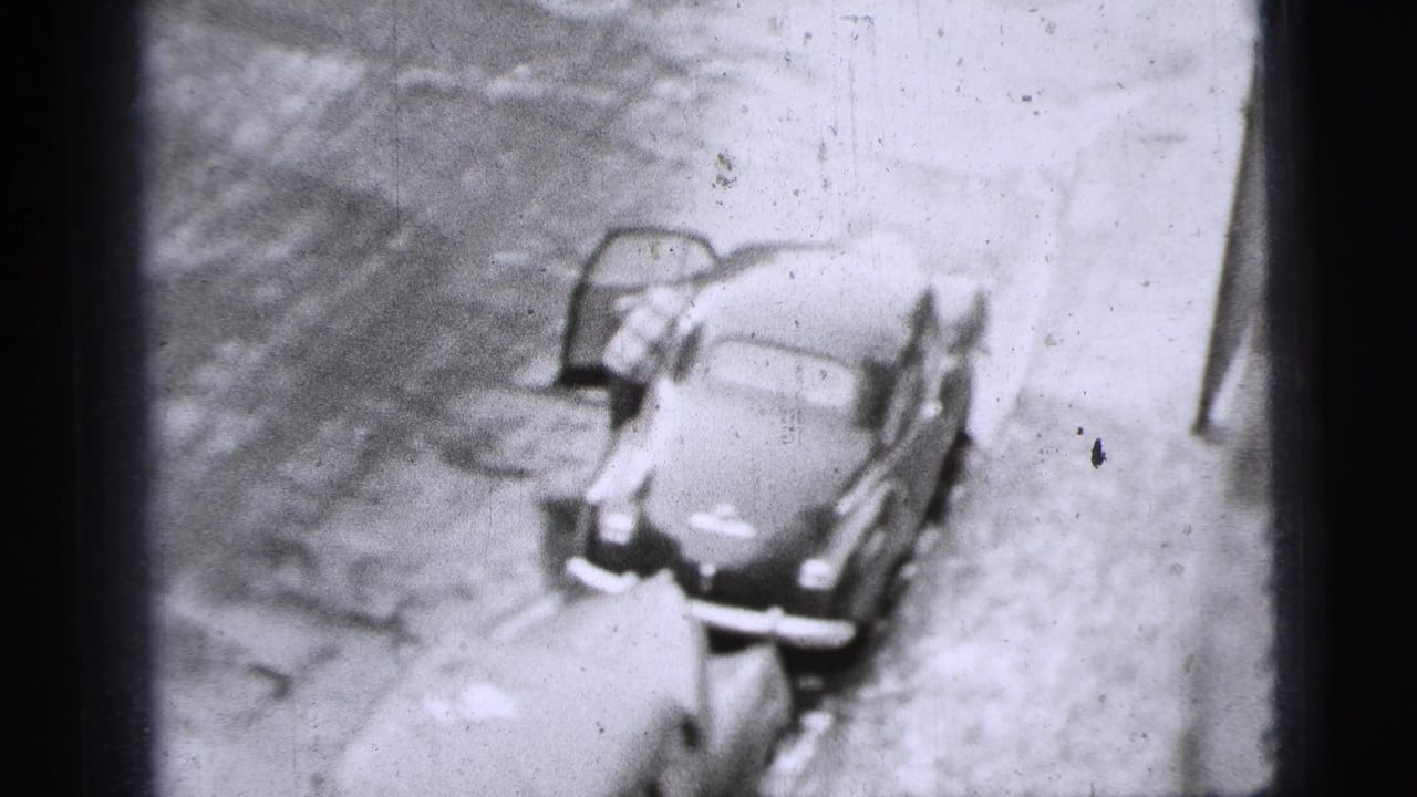High-angle 8mm view of a man at the driver's side of a parked mid-century car in New York City, with the door open.