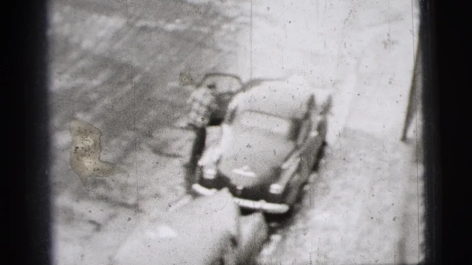 Soft 8mm frame of the man beside the open driver's door of the parked 1950s car on a New York City street.